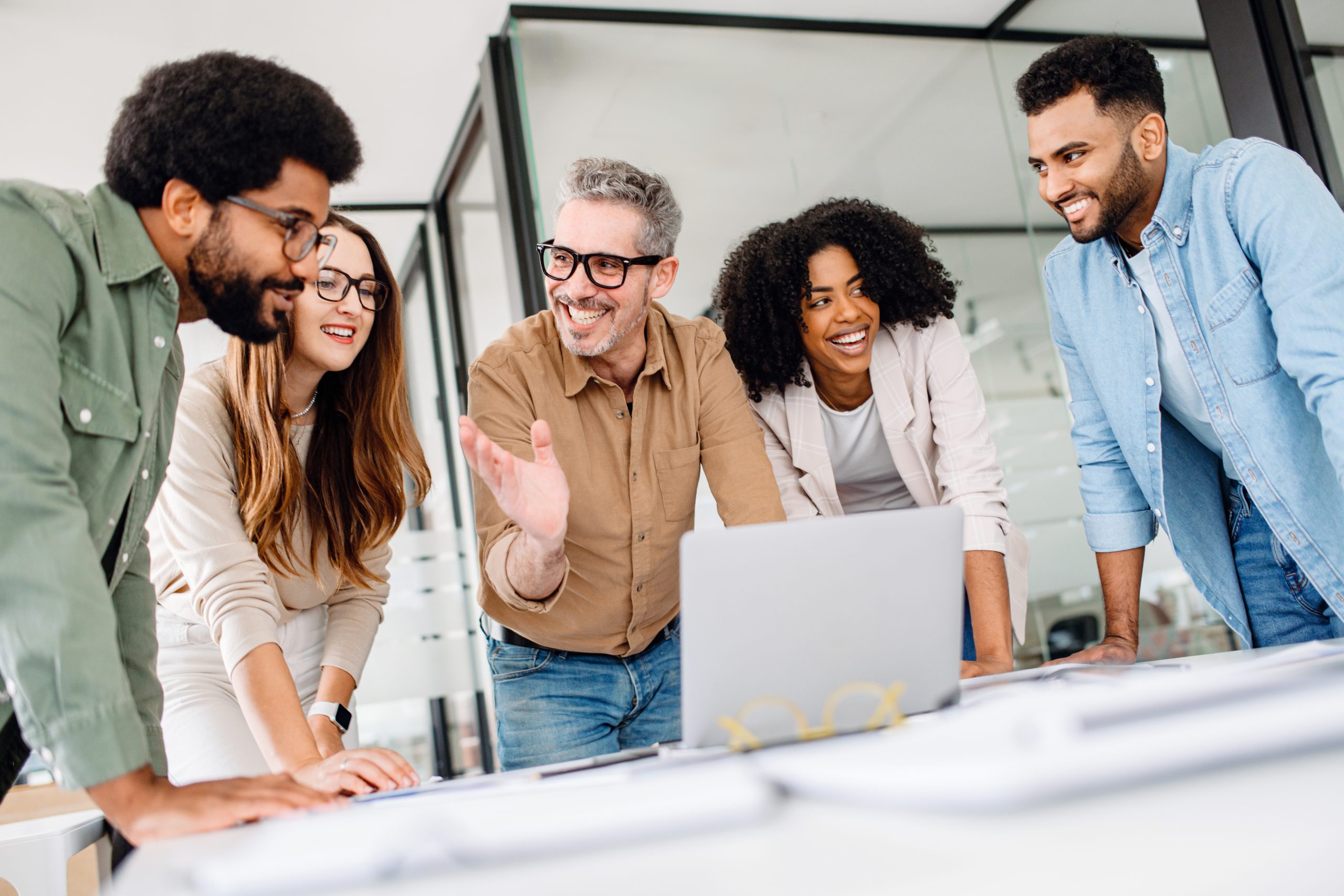 Diverse group of professionals leans in towards a laptop, sharing a moment of collaboration and interaction in their modern workspace. The energy and engagement of a diverse team working on a project