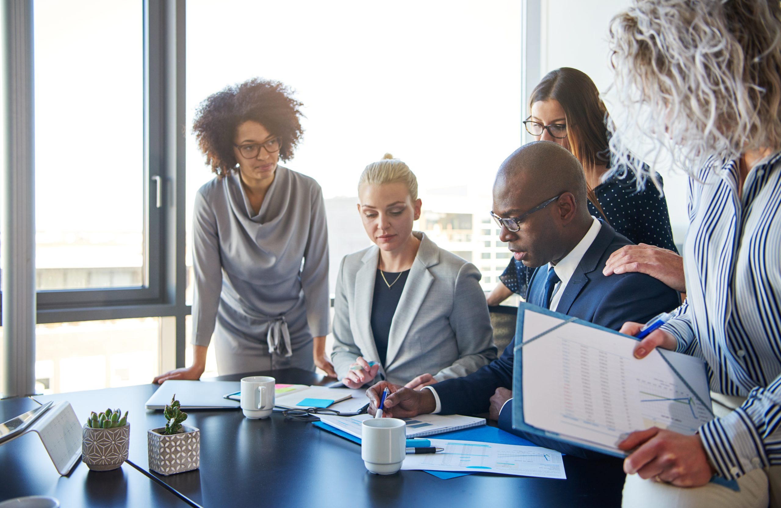 Group of focused businesspeople standing around their manager sitting at an office boardroom table reviewing charts and paperwork
